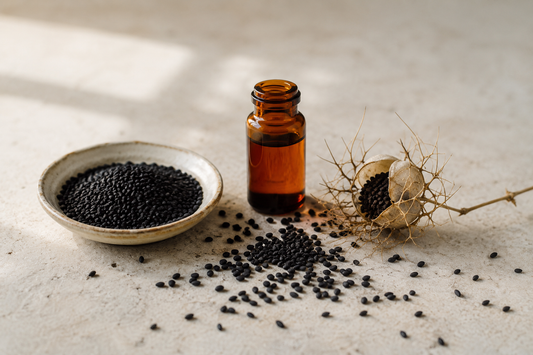 Black seed oil in a glass vial beside whole Nigella sativa seeds and a dried seed pod
